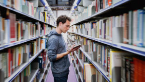 Person standing in between book shelves while looking at unopened book in hands