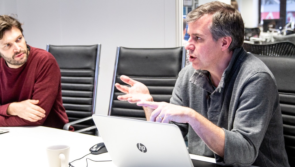 Two men having a discussion at a table, one with a laptop