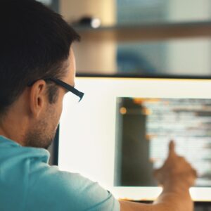 Rear view of partially unrecognizable man sitting in front of a computer pointing at a screen where are several lines of computer code.