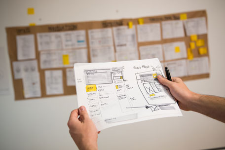 Pair of hands holding up a user interface sketch in front of a roll of brown paper on the wall, containing multiple other sketches