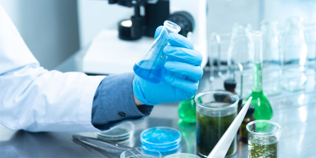 Close up of scientific equipment, including a beaker full of blue liquid being held in a gloved hand