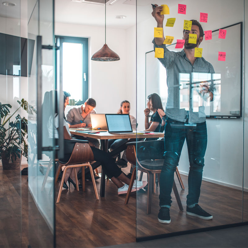 Meeting room with people at a table and one person sticking post-it notes on a glass wall